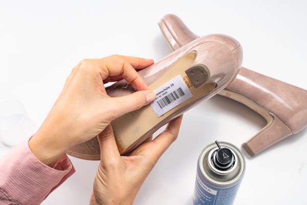 A hand holds a shoe with a price tag, while a spray can stands next to it. The shoe is a beige high heel.