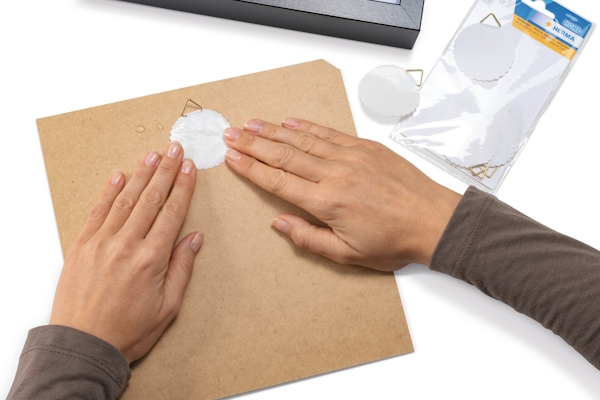 Hands are pressing a round hanger from HERMA onto a brown cardboard, while a pack of additional stickers lies next to it.