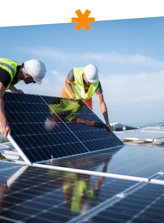 Two workers are installing solar panels on a roof, wearing safety helmets and reflective vests.
