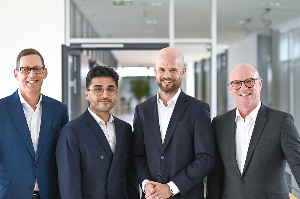 Four men in suits are standing in a modern office. They are smiling and posing together for a photo.