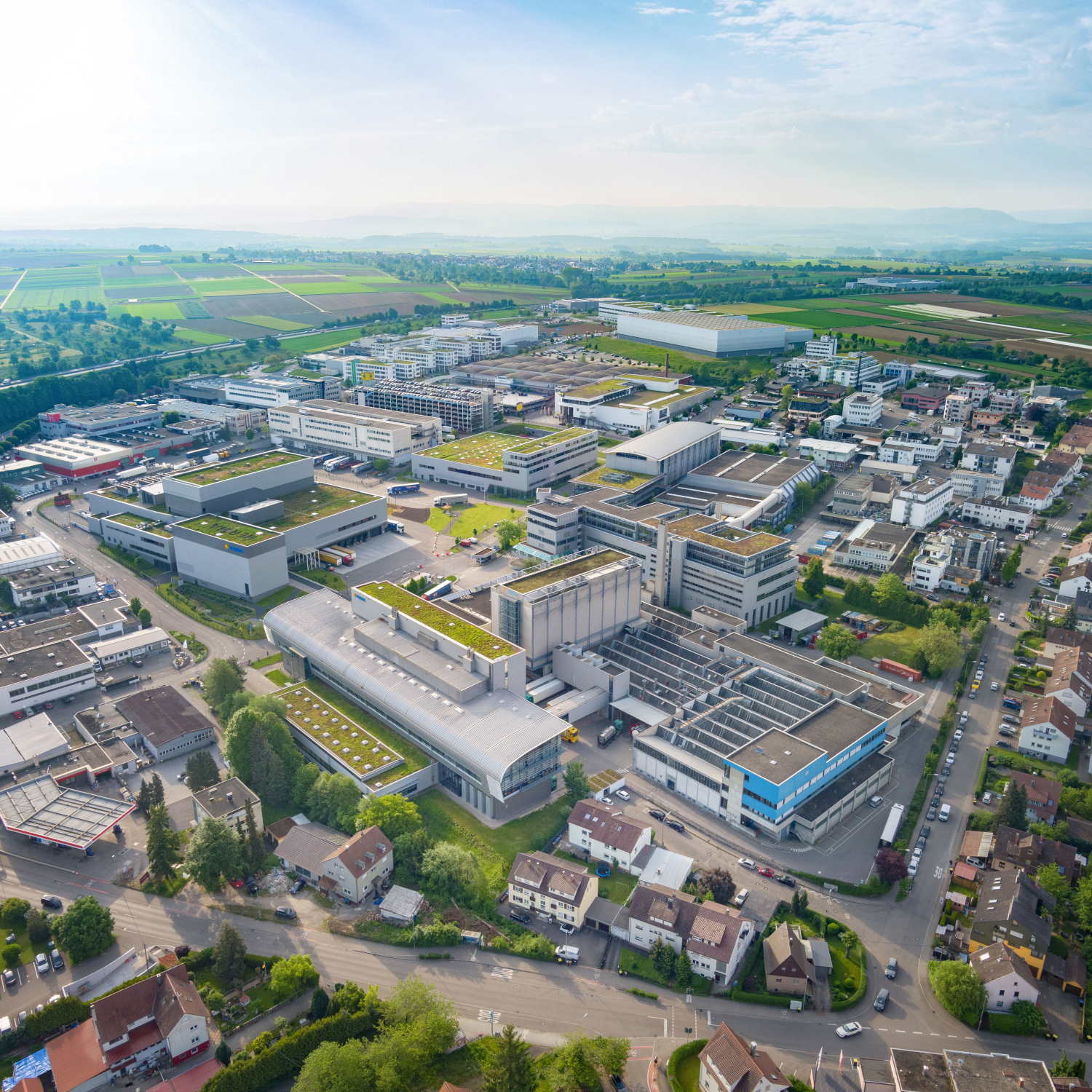 Luftaufnahme eines Industriegebiets mit modernen Gebäuden, Lagerhäusern und umliegenden Wohngebieten, umgeben von grünen Feldern.