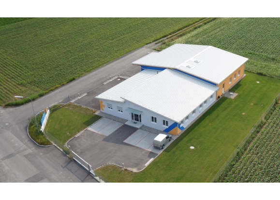 A modern, single-story building with a flat roof, surrounded by fields and a paved driveway.