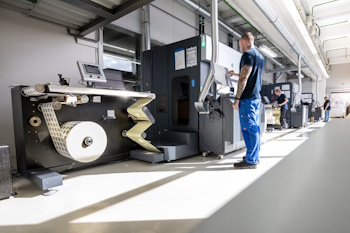 A worker operates a modern printing press in a production hall while a roll of paper is being prepared.