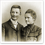 A man with a mustache and a woman in historical clothing pose together in a sepia-toned portrait.