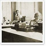 Three people are sitting at desks in an office, surrounded by typewriters and office supplies, with natural light coming in through the windows in the background.