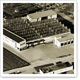 Aerial view of a large building with multiple roofs, surrounded by roads and fields. The building appears to have an industrial purpose.