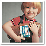 A smiling child with blonde hair is holding a football and wearing a red T-shirt with a black backpack.