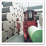 A woman stands next to a red forklift in a warehouse filled with large rolls of paper. She is holding a notebook and looking at it.