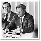 Two men are sitting at a table; one is speaking and gesturing while the other listens attentively. Both are dressed in formal attire.