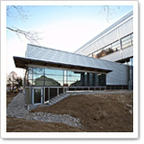 Modern buildings with glass facades and sloped roofs, surrounded by an unpaved area and trees in the background.