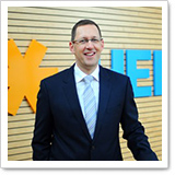 A man in a suit stands in front of a wooden wall with an orange and blue logo. He is smiling and appears professional.