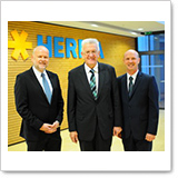 Three men are standing in a modern office with a wall displaying the word 'HERBA.' They are dressed in formal attire and smiling.