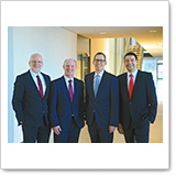 Four men in suits stand side by side in a modern office building. They are smiling and wearing ties in different colors.