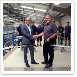 Two men shake hands in a factory hall, while several people stand in the background.