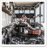 A digger is working in a demolished building, surrounded by debris and construction materials. Windows and walls have been partially removed.