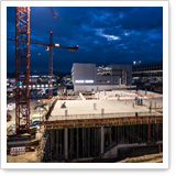 Construction site with a concrete foundation and cranes under a cloudy sky. Construction activities are visible, with building structures in the background.