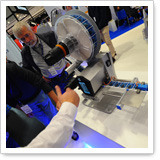 A person is demonstrating a technical device with a large, round component and a handle at a trade fair.