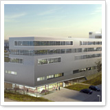 Modern, multi-story buildings with large windows and a bright facade, surrounded by green landscapes and parked cars.