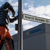 A worker is standing on a ladder installing a street sign that reads 'Heinrich-Hermann Street' in front of a company building.