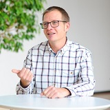 A man wearing glasses and a checkered shirt is sitting at a table, gesturing as he speaks. In the background, a plant is visible.