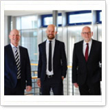 Three men in suits stand in a modern office building. They are smiling and posing amicably in front of large windows.