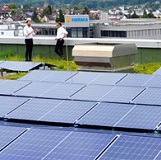Two people in suits are standing on a rooftop next to solar panels, having a conversation. Buildings are visible in the background.