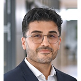 A man with dark, curly hair and glasses is wearing a suit and standing in a modern interior.
