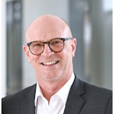 A man with glasses and a bald head is wearing a suit and smiling warmly. The background is blurred and bright.