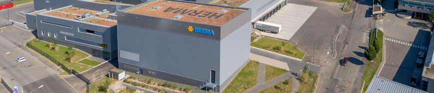 Modern warehouse with a green roof and the 'Hertila' logo on the facade, surrounded by parking lots and streets.