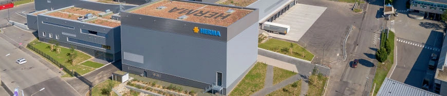 Modern warehouse with a green roof and the 'Hertila' logo on the facade, surrounded by parking lots and streets.