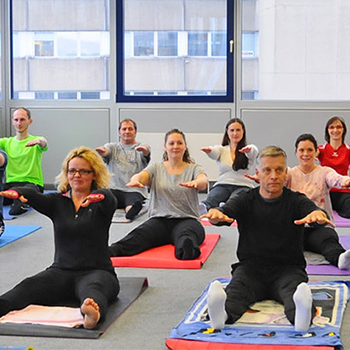 A group of people is practicing yoga in a room; some have their arms stretched out to the sides while others are sitting on mats.