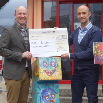 Two men are holding a large check for 10,000 euros. In the background, colorful painted figures can be seen.