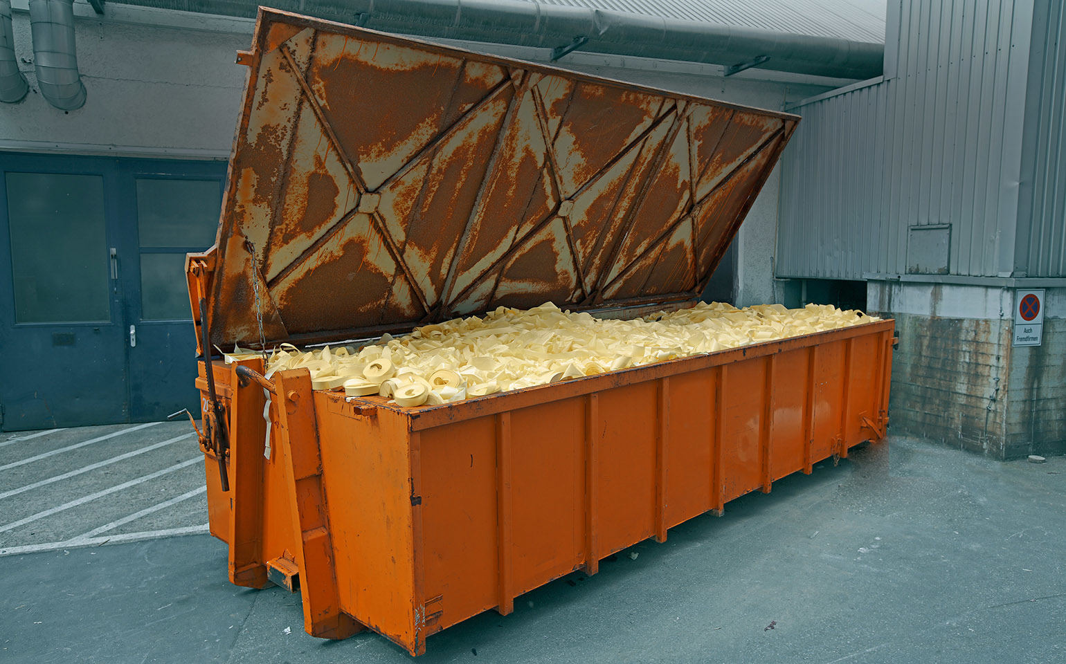 A large orange container filled with yellow waste stands against a wall, with its lid open.