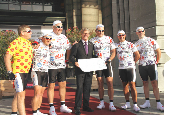 Six cyclists in jerseys are standing with a man in a suit and tie, who is holding a large sign, in front of a building.