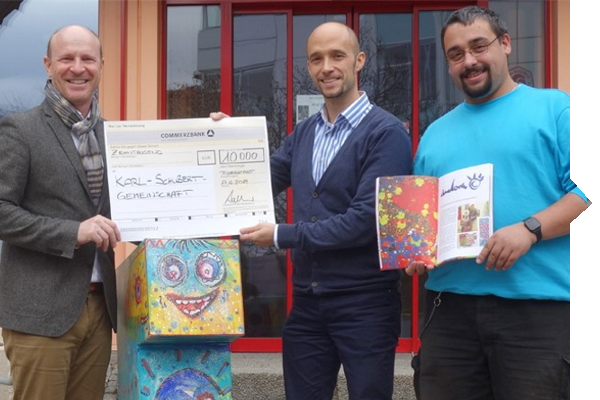 Three men are holding a large check for 10,000 euros and a colorful art book. In the background, a vibrant piece of artwork can be seen.
