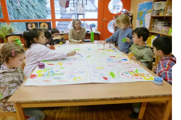 Children are painting together at a large table while an adult guides them. Colorful drawings and art supplies are visible.