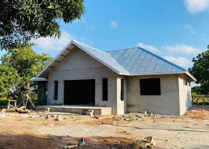 A half-finished house with a blue roof, large windows, and a concrete foundation, surrounded by unpaved ground and trees.