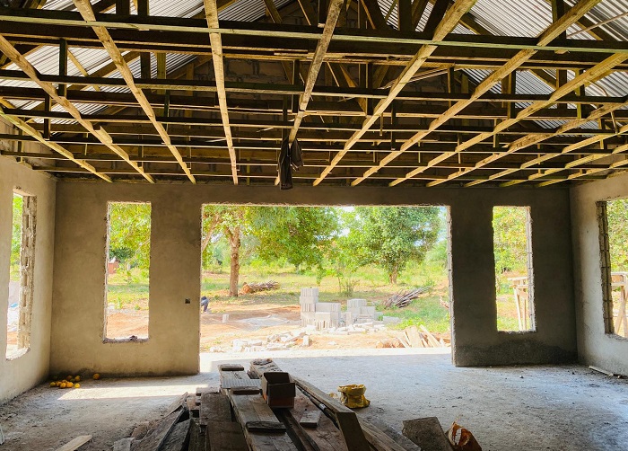 An unfinished space with exposed rafters and large windows overlooking a green outdoor area. Wooden materials are scattered on the floor.
