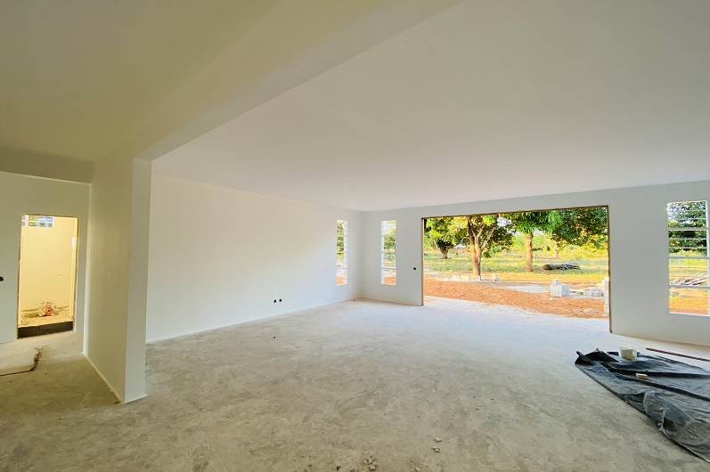 Bright, empty room with white walls and large windows overlooking a garden. The floor is unfinished.