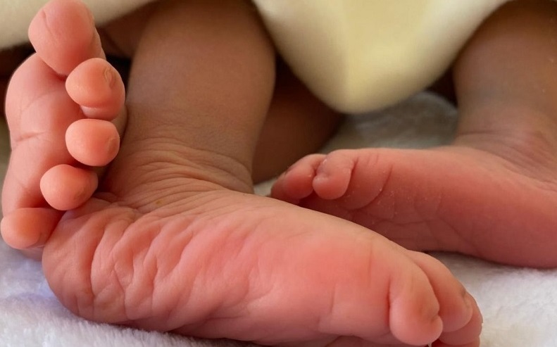 Two tiny baby feet rest on a soft surface, partially covered by a blanket. The skin is delicate and rosy.