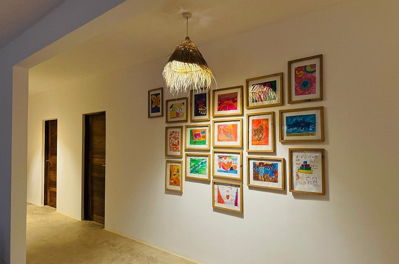 A hallway with a wall decorated with framed children's drawings. Two doors are visible in the background. A hanging lamp is also present.