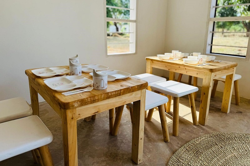 Two wooden tables set with dishes and cups, surrounded by chairs, in a bright room with windows and natural light.
