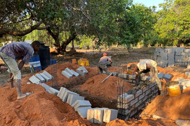 Workers are building with bricks and concrete in a construction area, surrounded by soil and trees.
