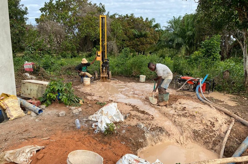 Two workers are drilling a hole in the ground, while a drill stands in the background, surrounded by dirt and water.