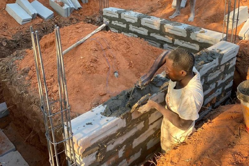 A worker is laying cement blocks at a construction site to build a wall. The ground is covered with red sand.