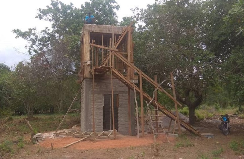 An unfinished building with a wooden frame and stairs, surrounded by trees and grass. A person is working on the roof.