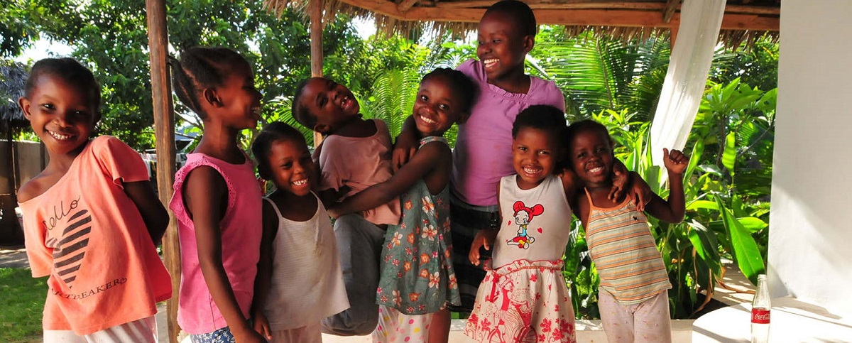 A group of eight girls stands together, smiling and happily posing in a green, sunny setting.