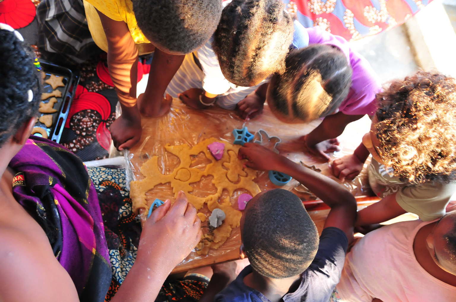 Children are shaping cookies from dough using cookie cutters on a table. Some kids are actively participating while others are watching.