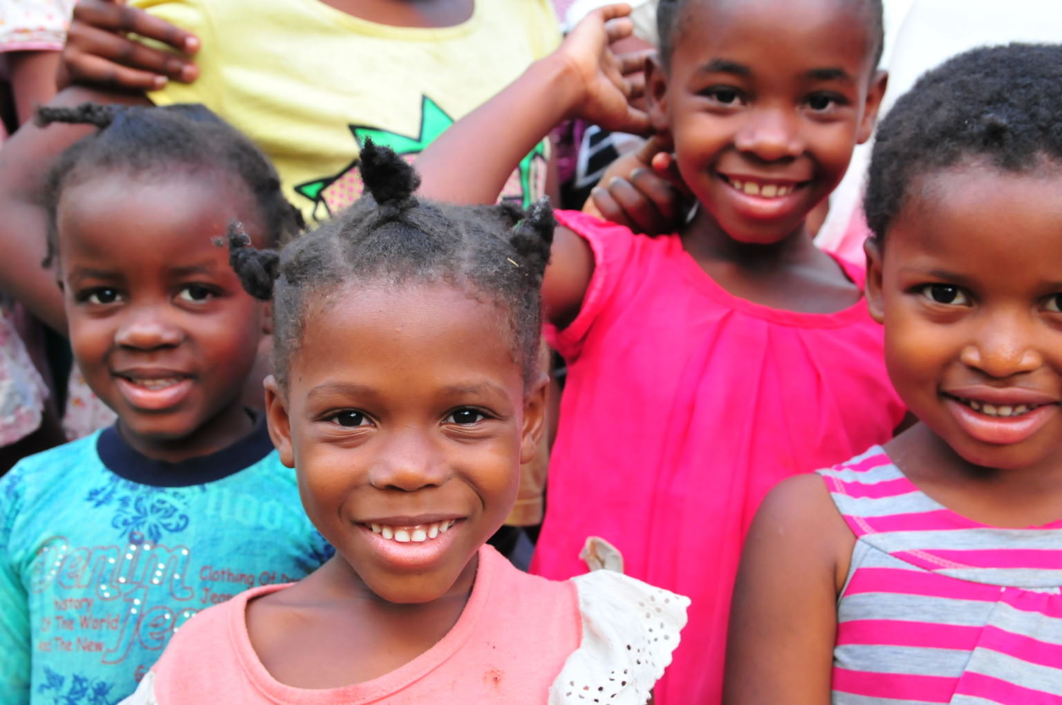 Four smiling children stand close together, wearing colorful clothes and radiating joy.