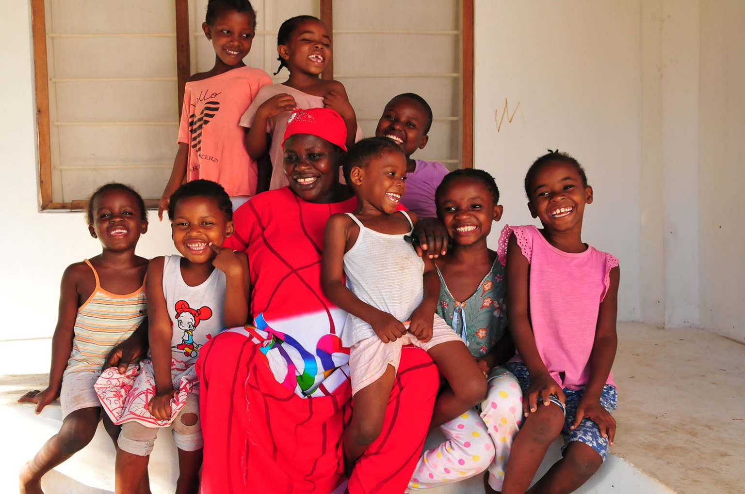 A group of children and an adult are sitting together and smiling. The children are wearing various colorful outfits.
